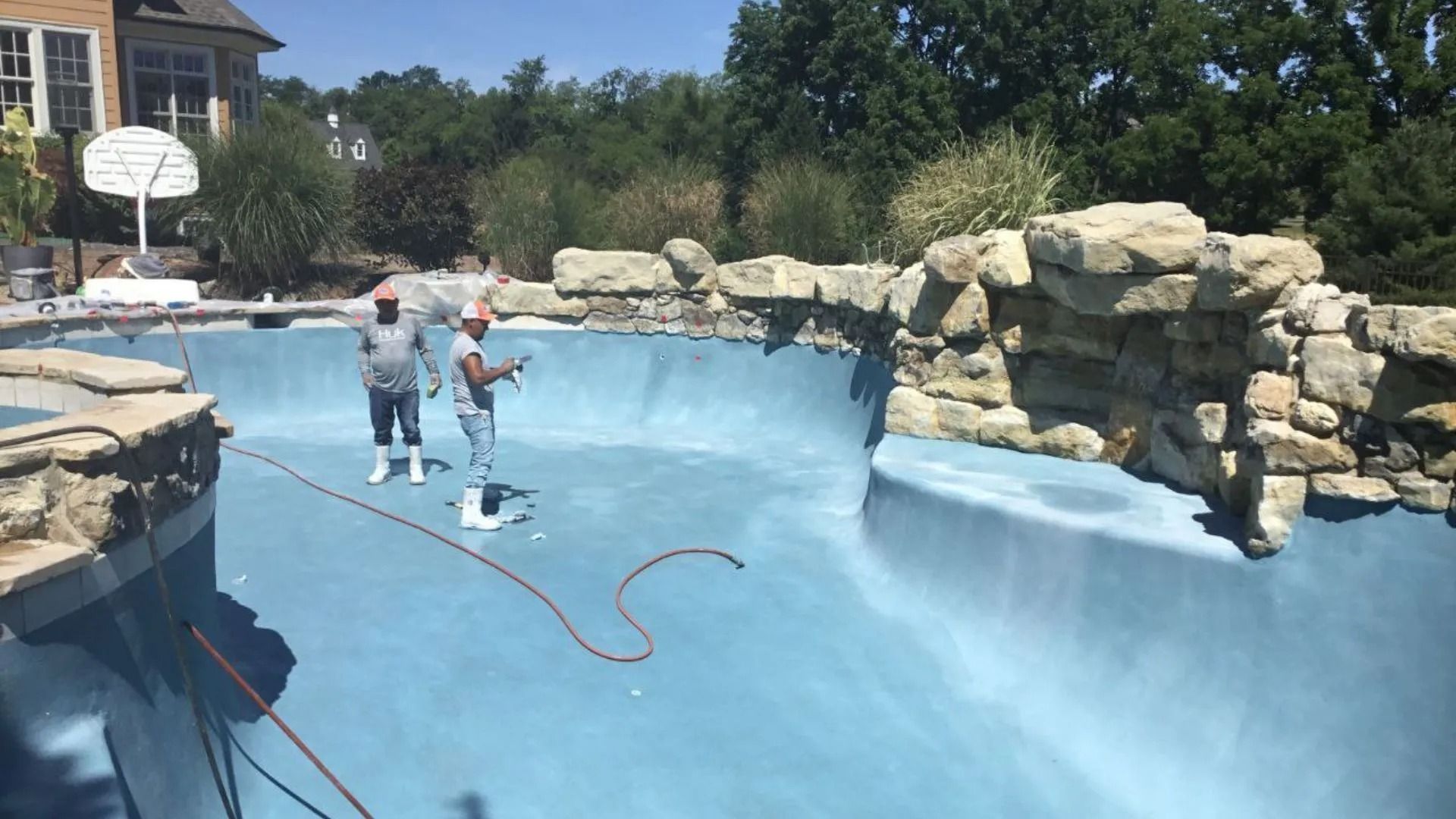 Two people are inside a light blue, empty pool, possibly cleaning or repairing it, with a rocky waterfall feature.
