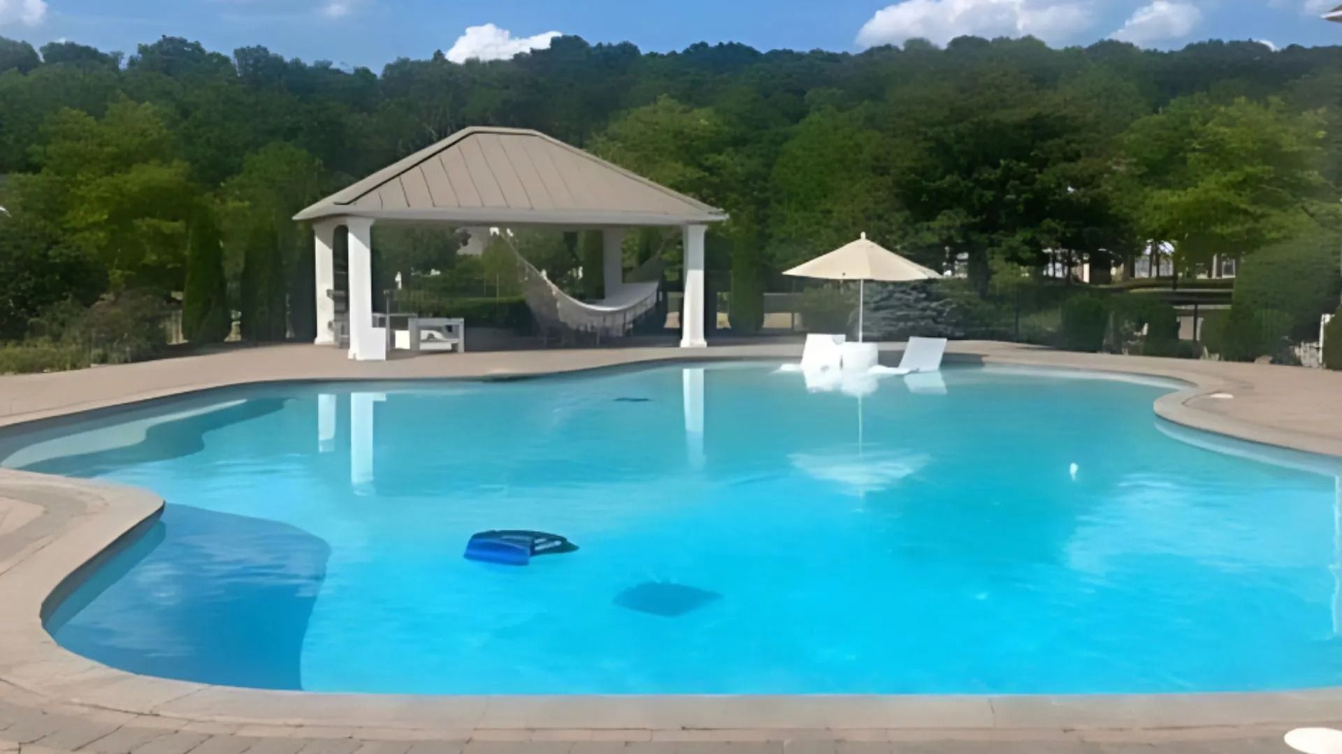 Pool with a gazebo, umbrella, and hammock. Turquoise water, surrounding trees, and clear sky.