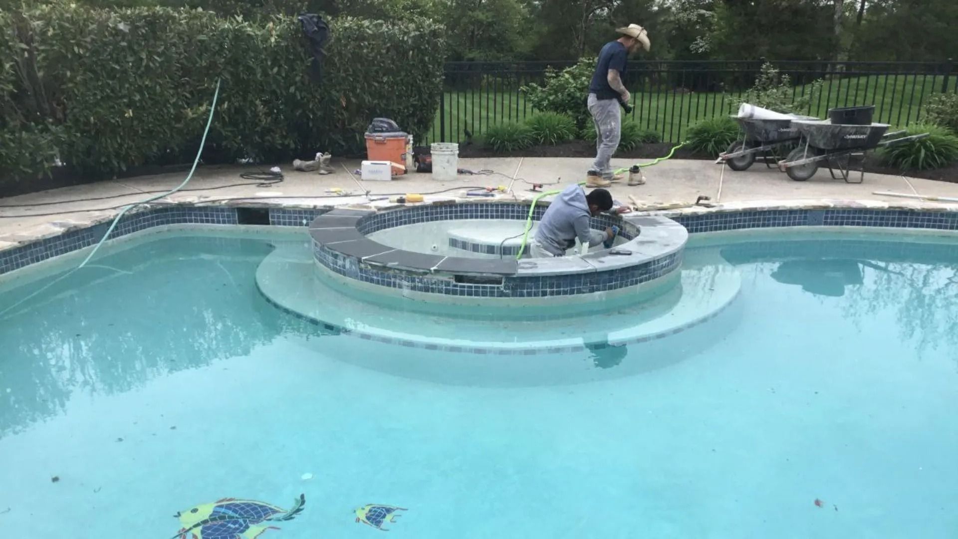 Workers tiling a pool's circular fountain. Blue water, tools, and a surrounding stone patio are visible.