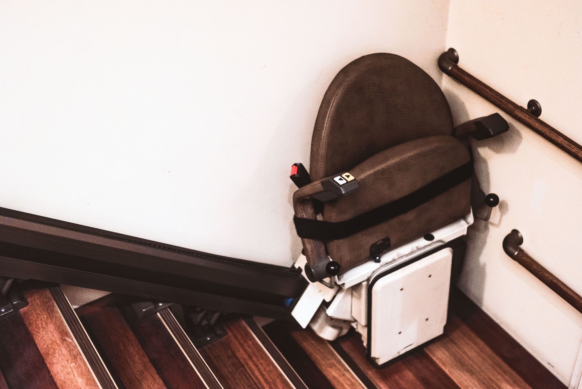 A stair lift chair folded against a white wall and wooden stairs, brown chair, handrails.