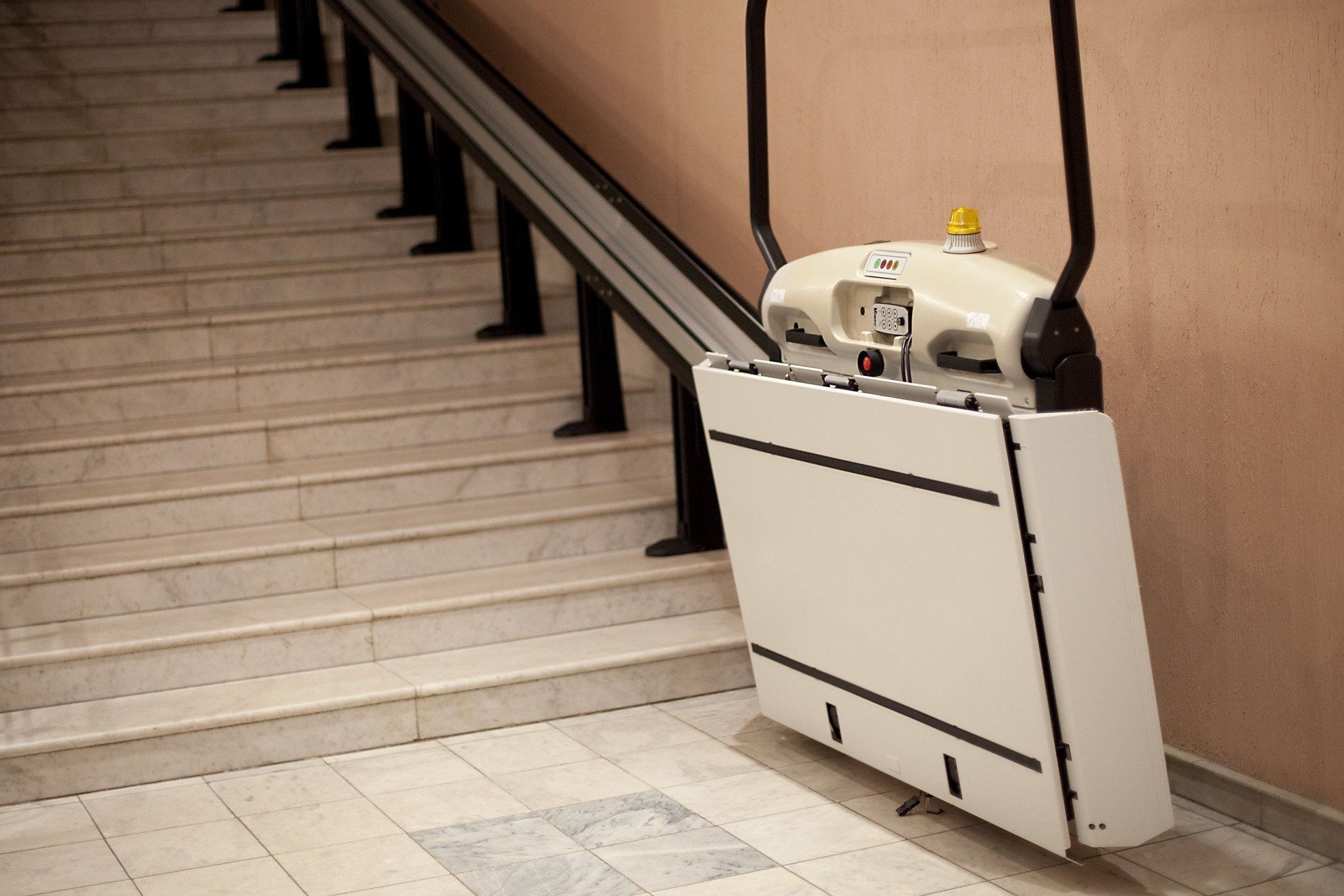 Stair lift on a staircase. Beige lift car travels along a black rail. Beige and white steps.