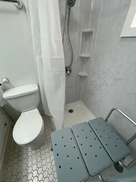 Bathroom with a walk-in shower, toilet, and a shower chair. White and gray tones.