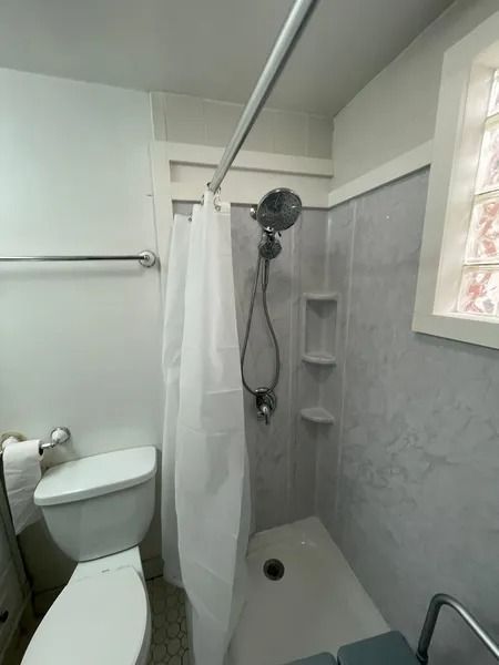 Small bathroom with toilet and shower. White shower curtain, gray tiled walls. Window on right.