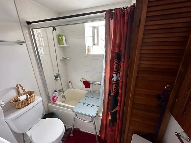 Bathroom with tub, shower curtain, toilet, and wooden door. A shower chair sits in front of the tub.