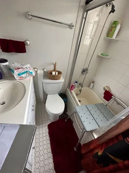 Small bathroom with a toilet, sink, and tub. A shower chair is near the tub.