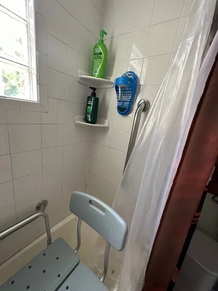 Shower with safety features: grab bar, shower chair, and corner shelves holding toiletries. White tiles.