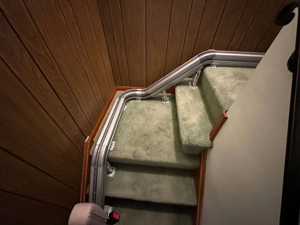 Staircase with a stairlift. Green carpeted steps, wood paneling, metal handrails.