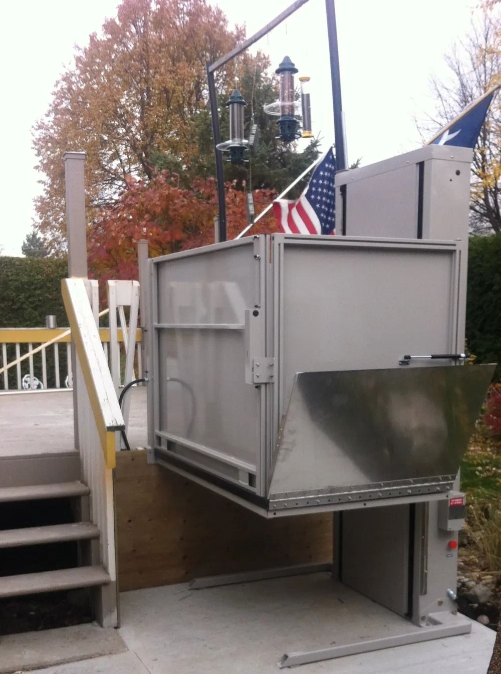 Outdoor wheelchair lift on deck with stairs, American flag, autumn trees.