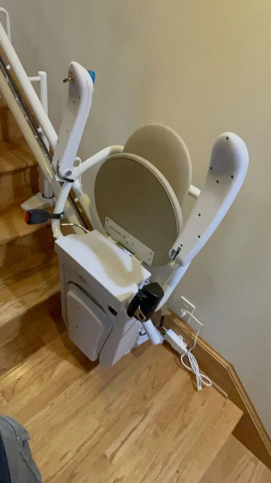 Stair lift on wooden stairs, folded up against a beige wall. White chair, tan seat cushions.