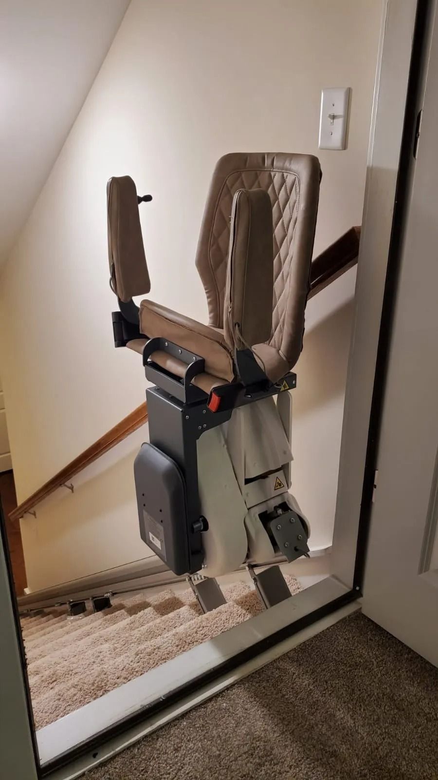 Stairlift installed on a staircase with beige seat and arms, against a beige wall and carpeted stairs.