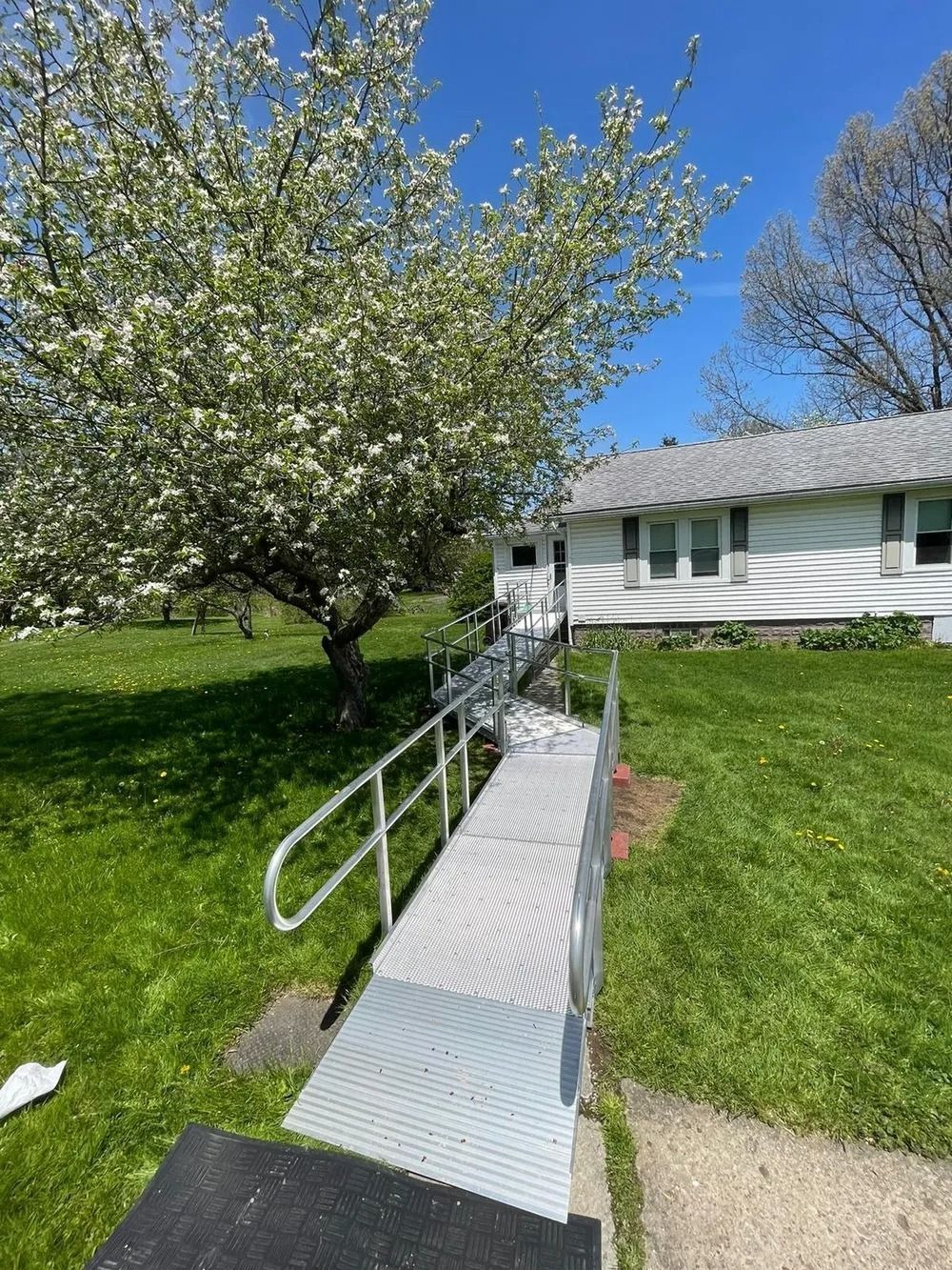 Metal ramp leads to white building with blooming tree, green grass, and blue sky.