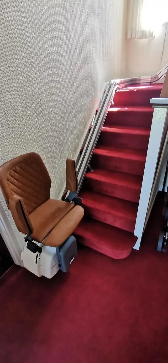 A stairlift with a brown seat beside a staircase covered in red carpet. The wall is textured.