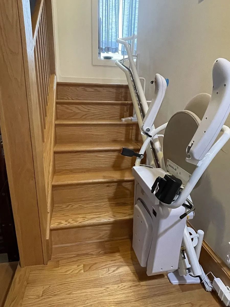 Stair lift on wooden stairs; beige seat, white frame. Staircase by wall with window.