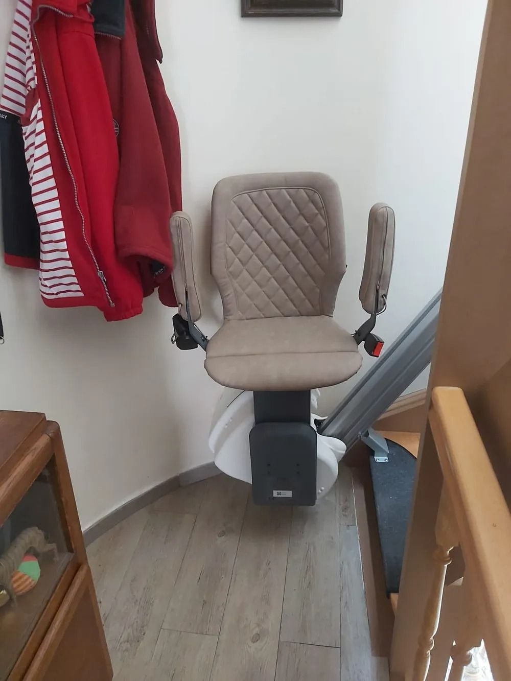 Beige stair lift chair on curved staircase. Coats hanging on wall.