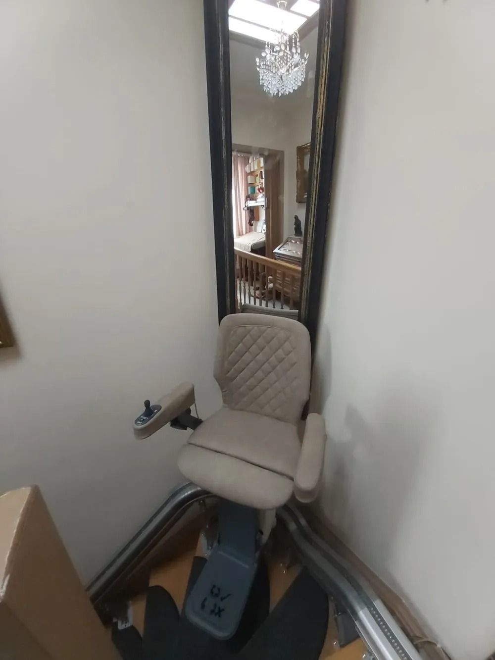 Stairlift chair in a corner, with reflective mirror showing a chandelier, a stairwell, and a glimpse of a room.