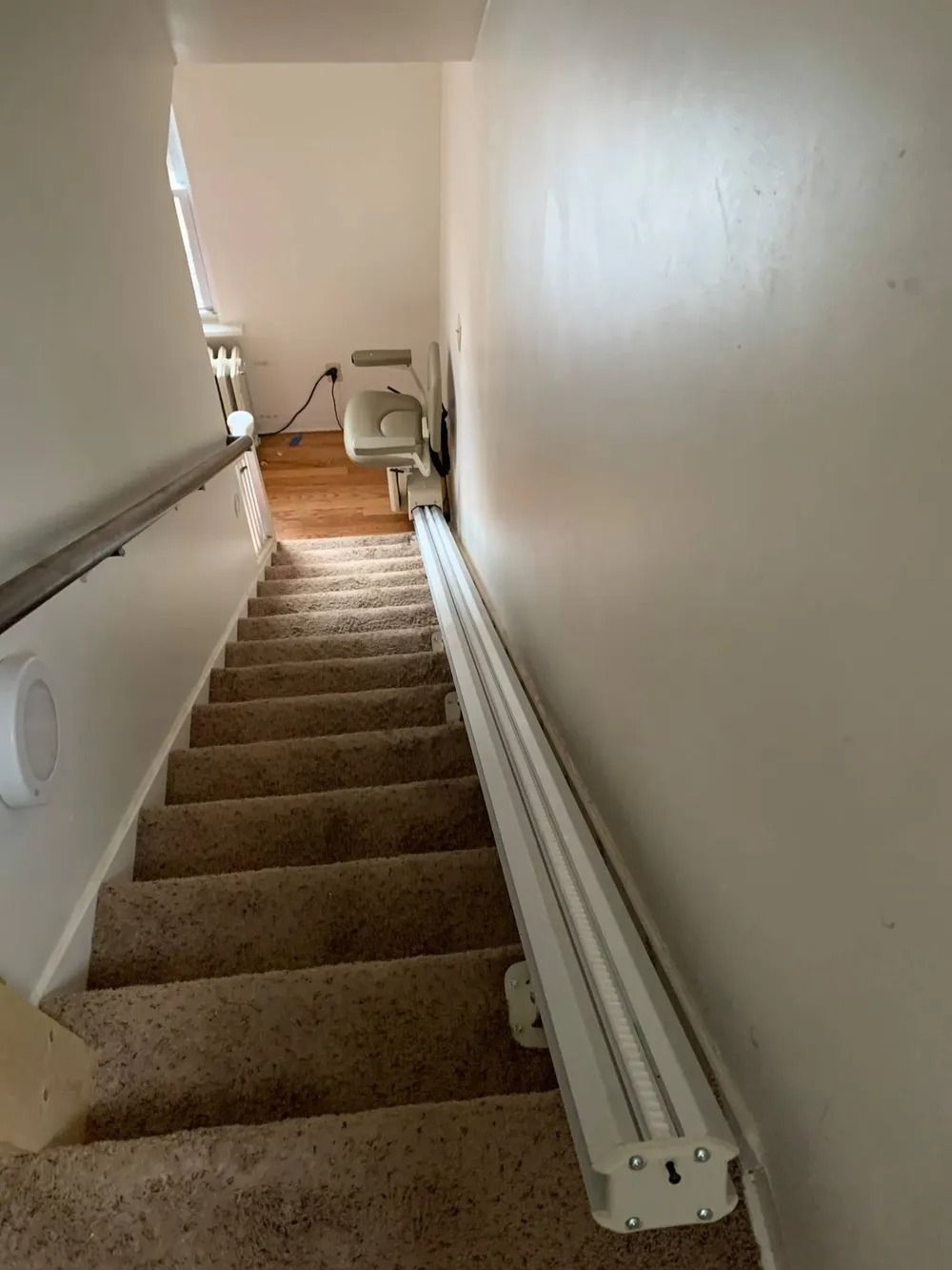 Staircase with a chair lift installed on the side; cream walls and tan carpet.