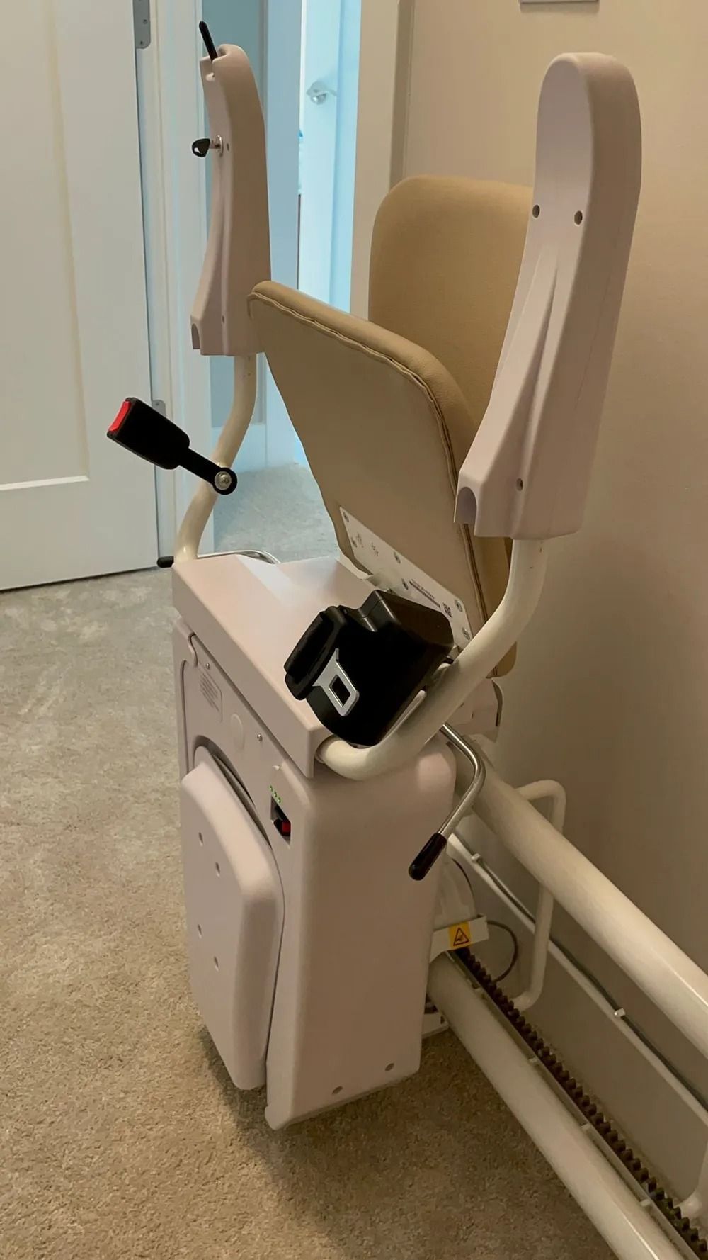 Stair lift mounted on a wall in a home. The seat is beige with a black seatbelt.