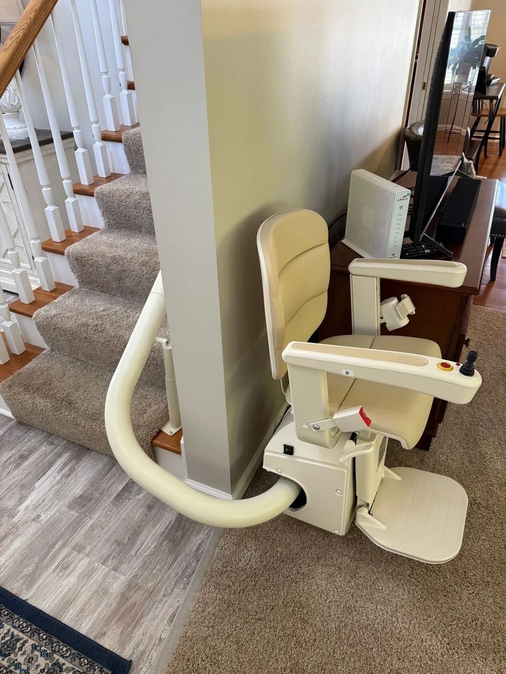 A beige stairlift is installed next to a staircase. The lift is against a gray wall and brown carpet.