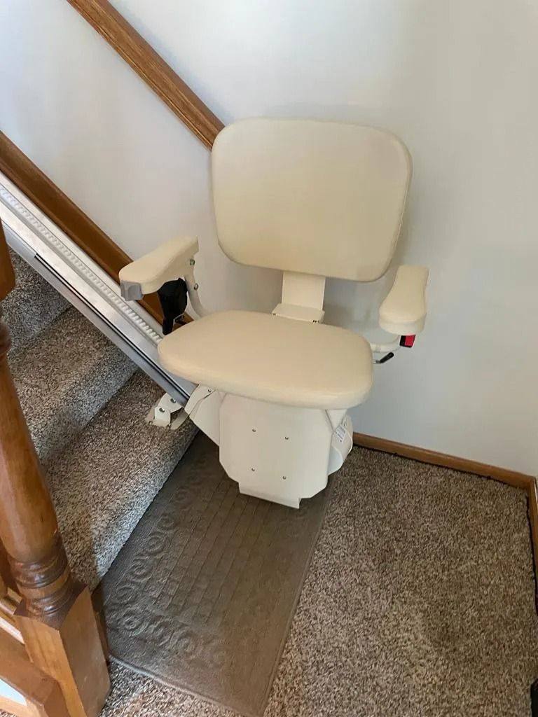Stair lift chair, beige, mounted on a staircase with tan carpet, brown handrail, against a white wall.