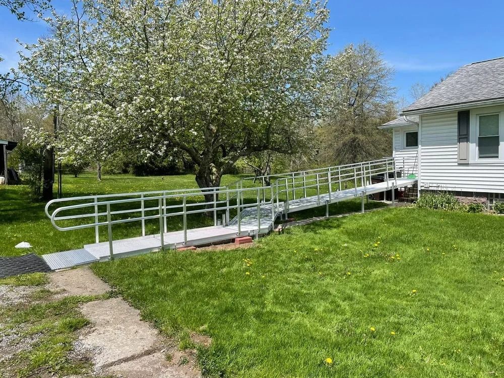 A metal ramp with handrails leads to a white building; a large tree with white blossoms is in the background.