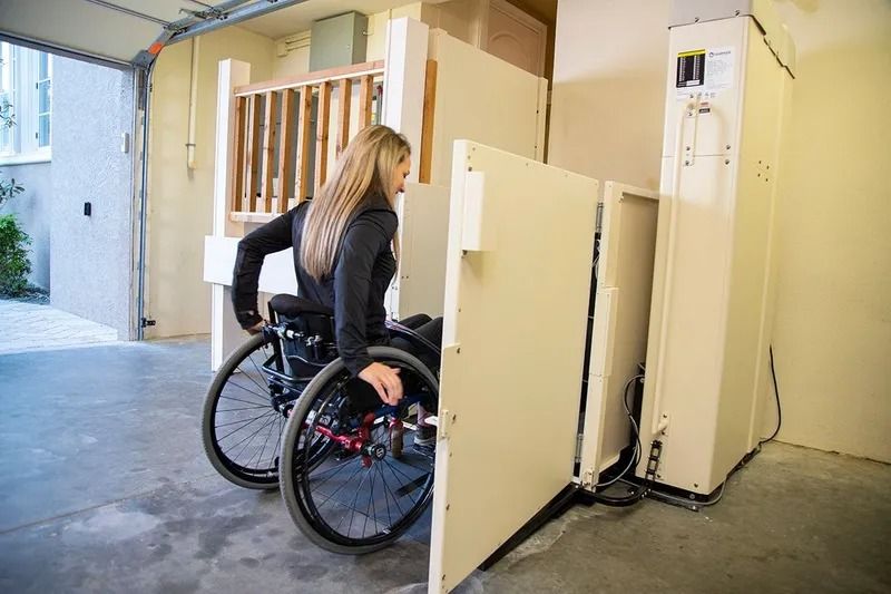 Woman in wheelchair using a vertical platform lift in a garage to access a higher level. Beige lift, open door.