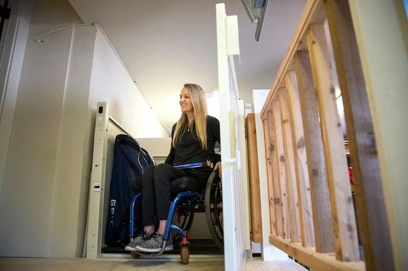 Woman in wheelchair using a vertical platform lift inside a home. Light-colored walls and wood stairs nearby.