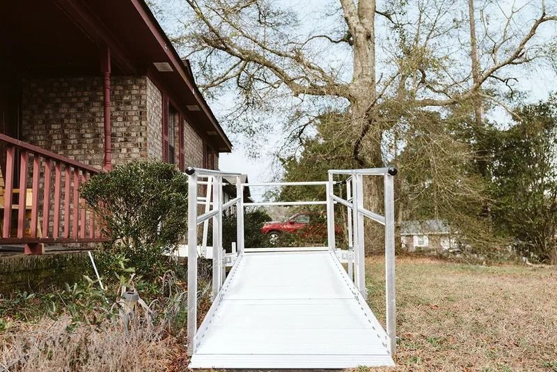 Aluminum wheelchair ramp leading to a brick house with a porch, set in a yard with trees.