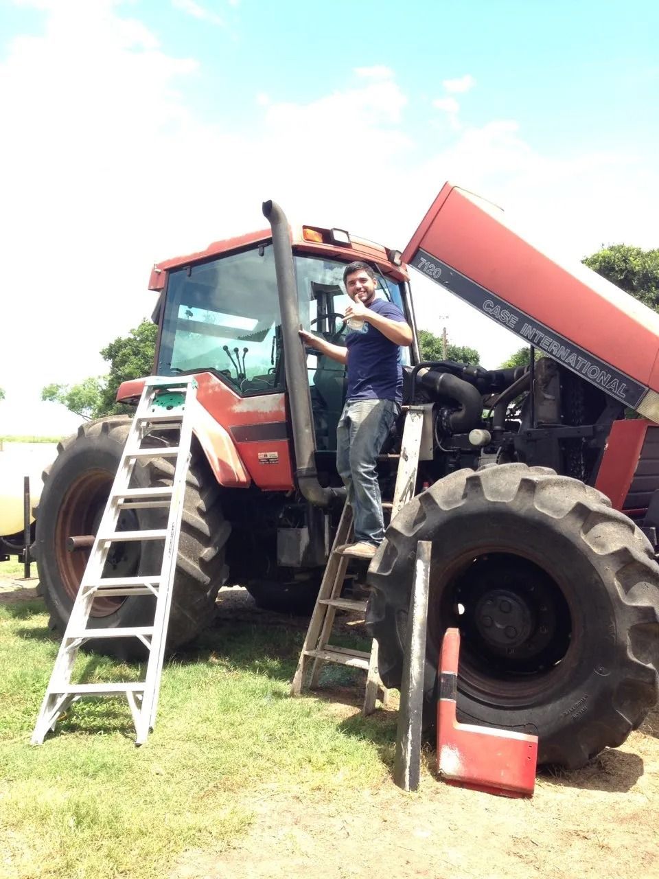 Man standing on a tractor, holding a tool near the exhaust pipe; open hood, ladder.