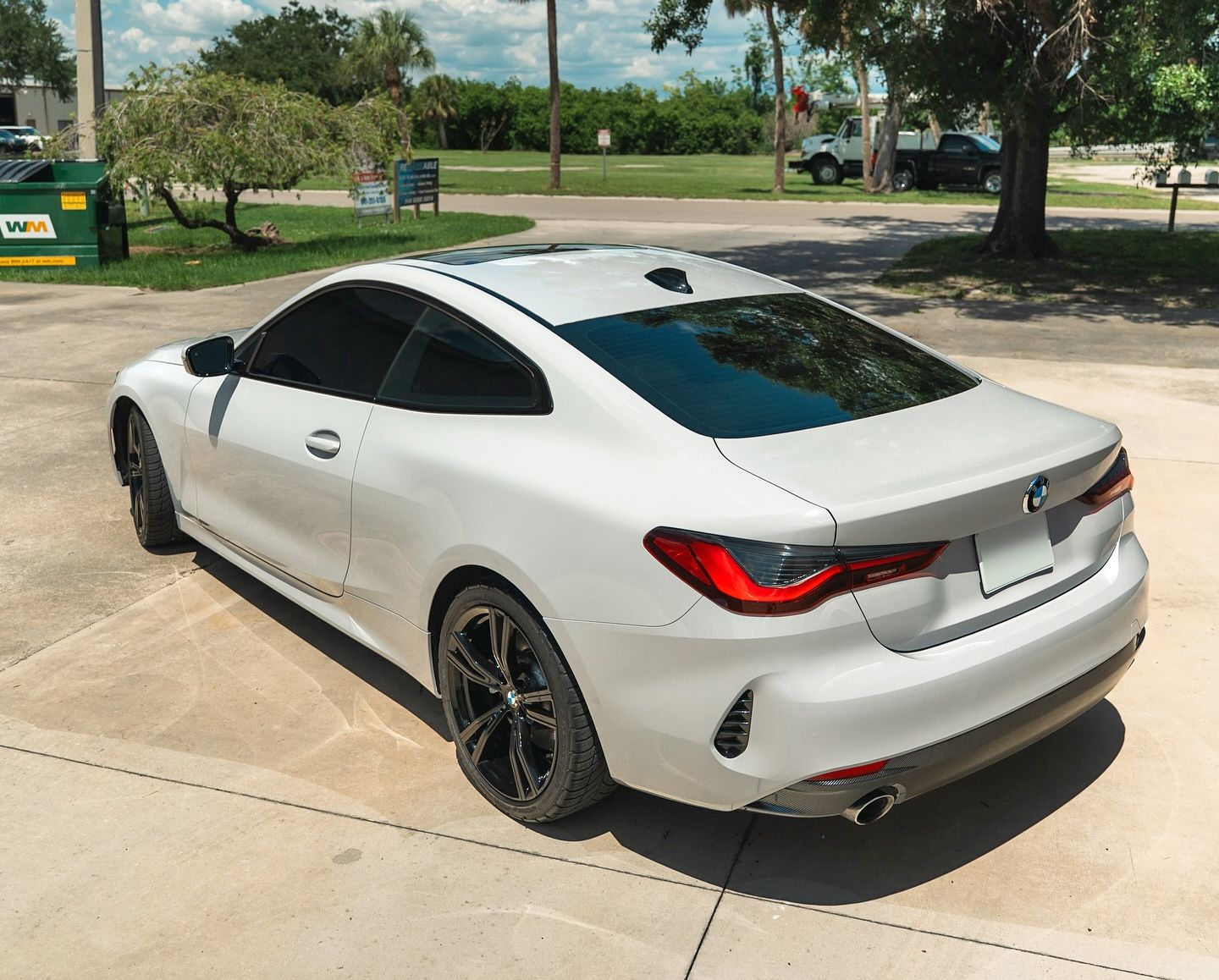 White BMW coupe parked on a paved driveway with a green dumpster in the background under a sunny sky.