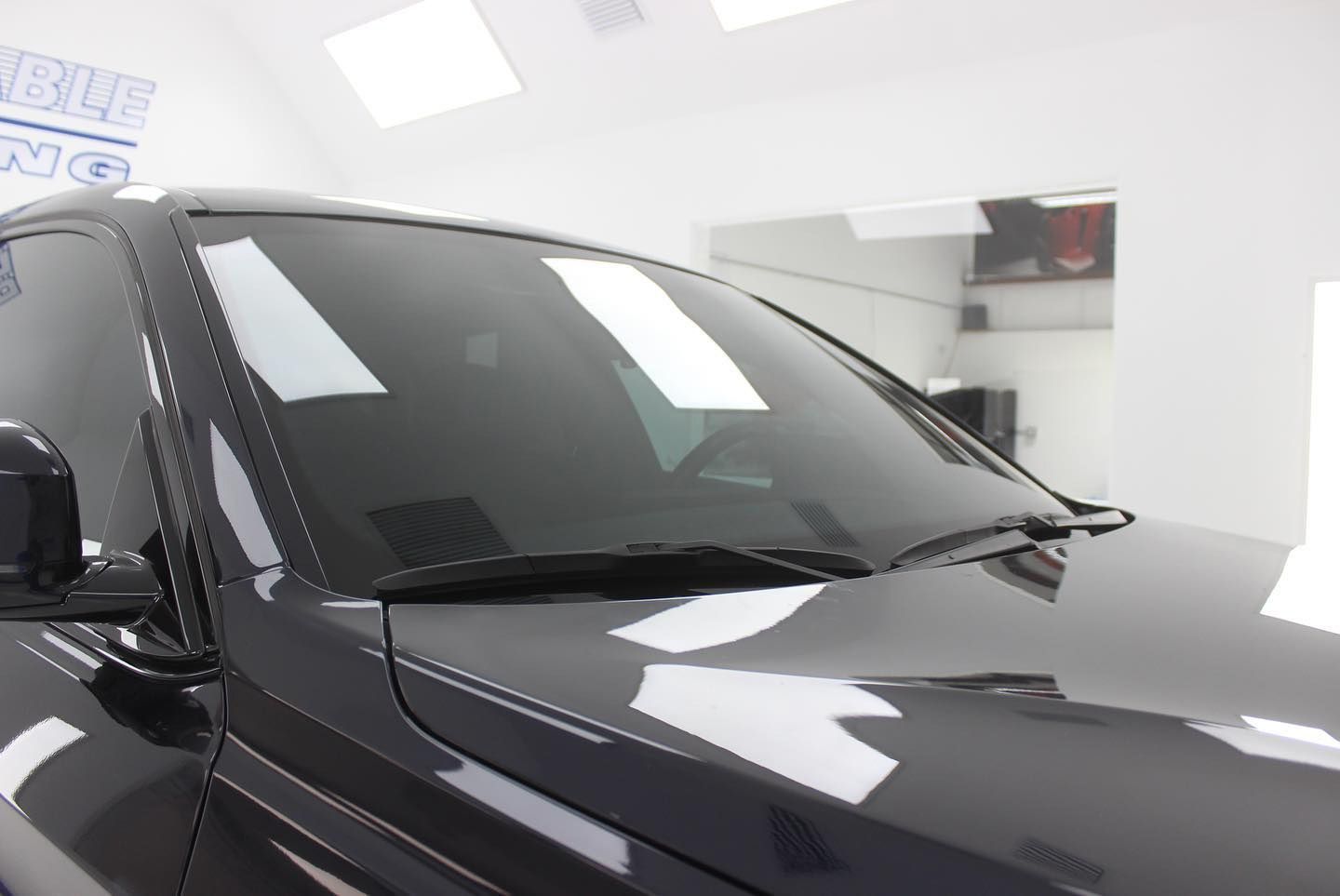 Dark-colored car with tinted windows in a white-walled room; the windshield reflects the room's lights.
