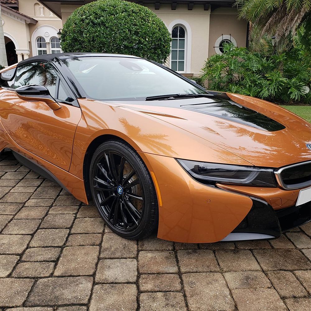 Orange and black BMW i8 sports car parked on a brick driveway, house in the background.