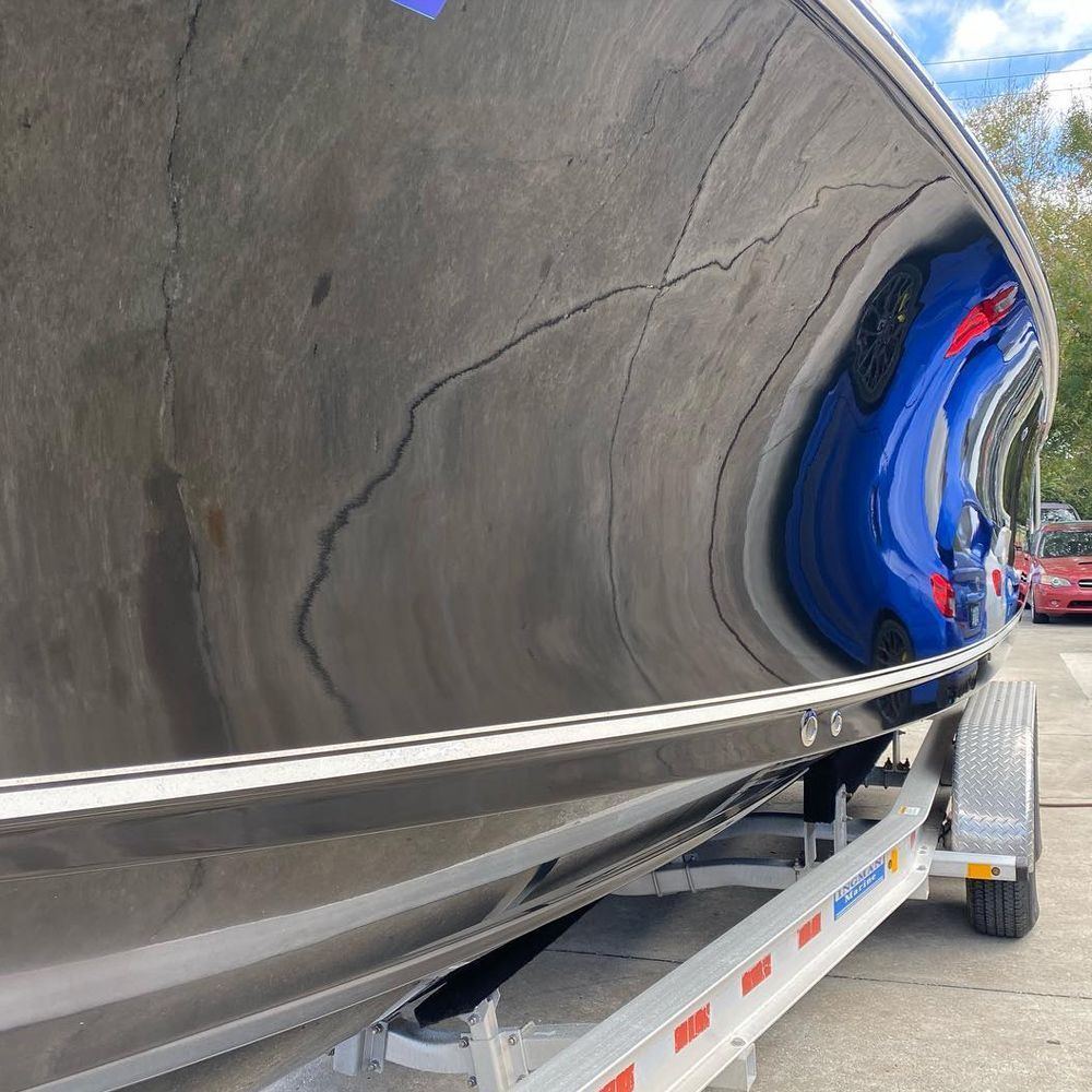 Black boat on a trailer, reflecting a blue car. Sunny outdoor setting.