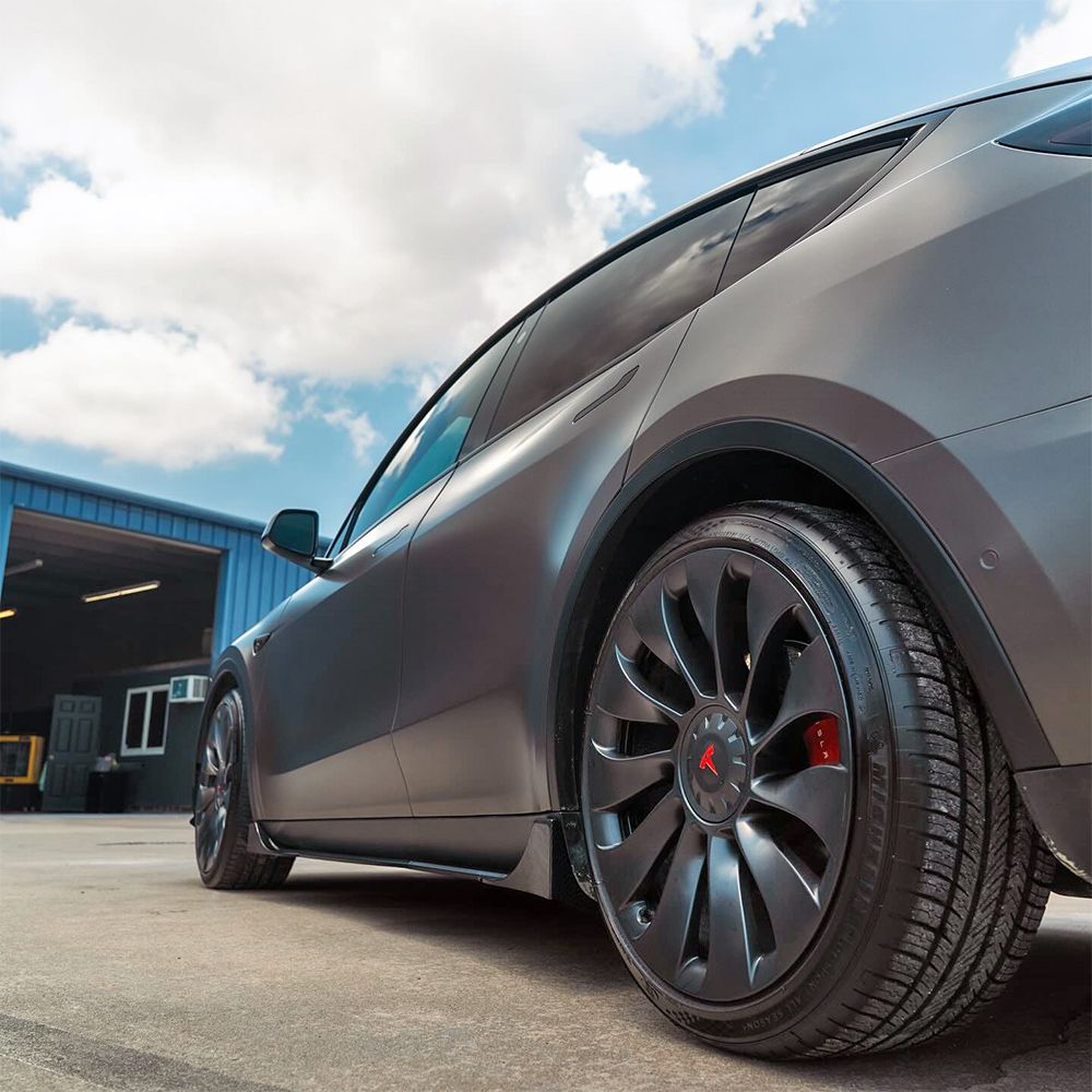 Gray Tesla SUV with black rims parked outside a blue garage, against a cloudy sky.