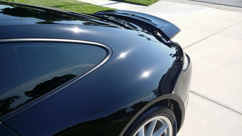 Black car's glossy rear section, with a spoiler and a window, parked on a paved driveway in sunlight.