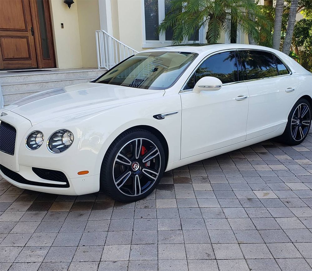 White Bentley sedan parked on a paved driveway in front of a house.