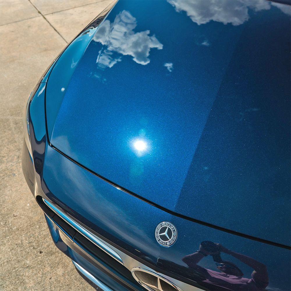 Blue car hood with Mercedes logo; reflection of clouds and sunlight.