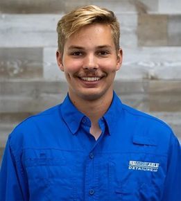 Smiling man in a blue button-up shirt with logo, in front of a wood-paneled wall.