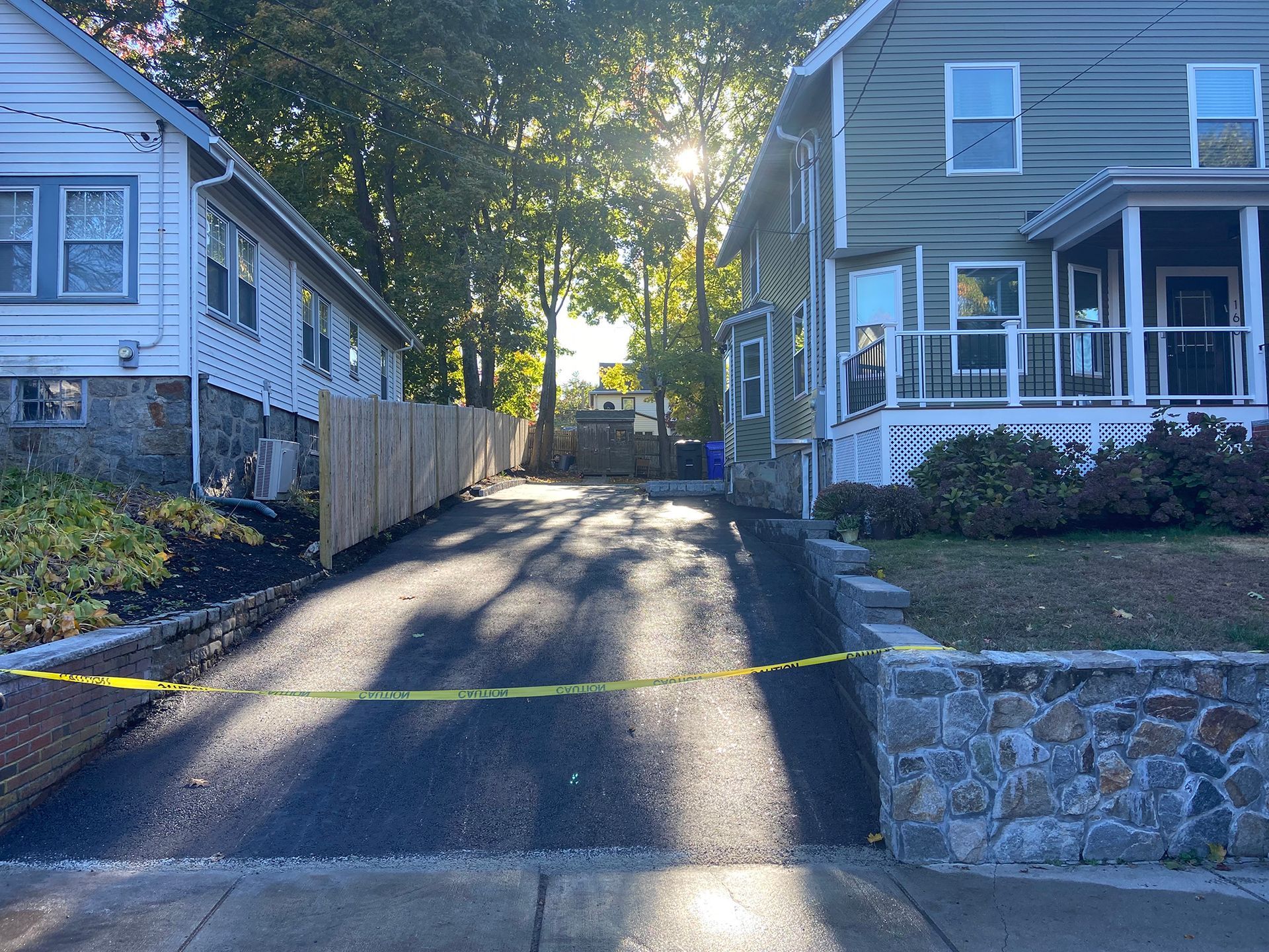 A paved driveway between two houses, blocked by yellow caution tape, with trees and sunlight in the background.