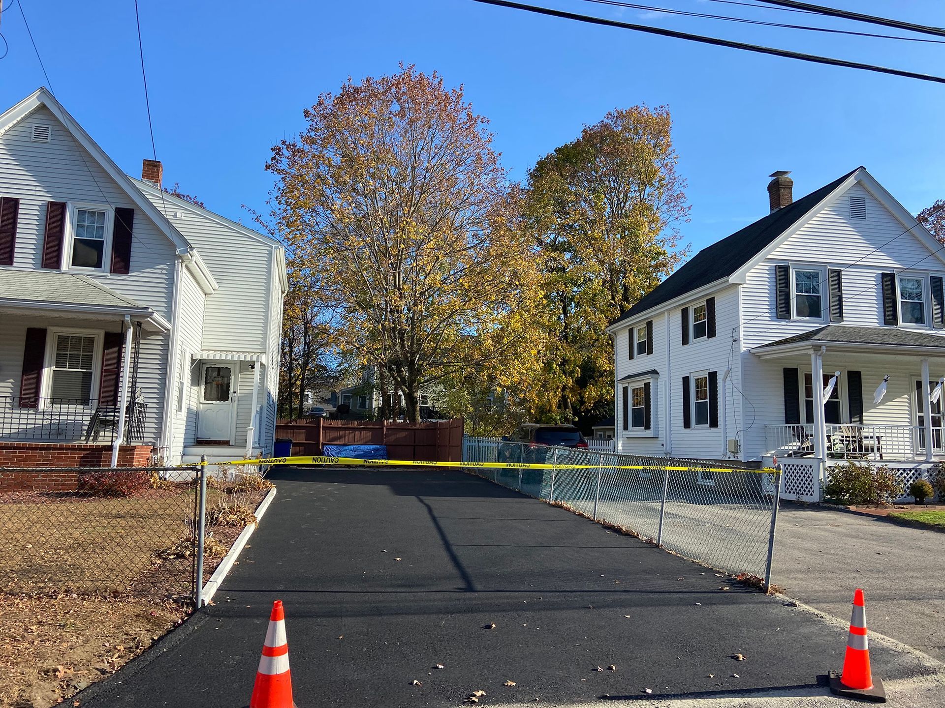 Two white houses separated by a newly paved black driveway, marked with orange traffic cones and yellow caution tape.