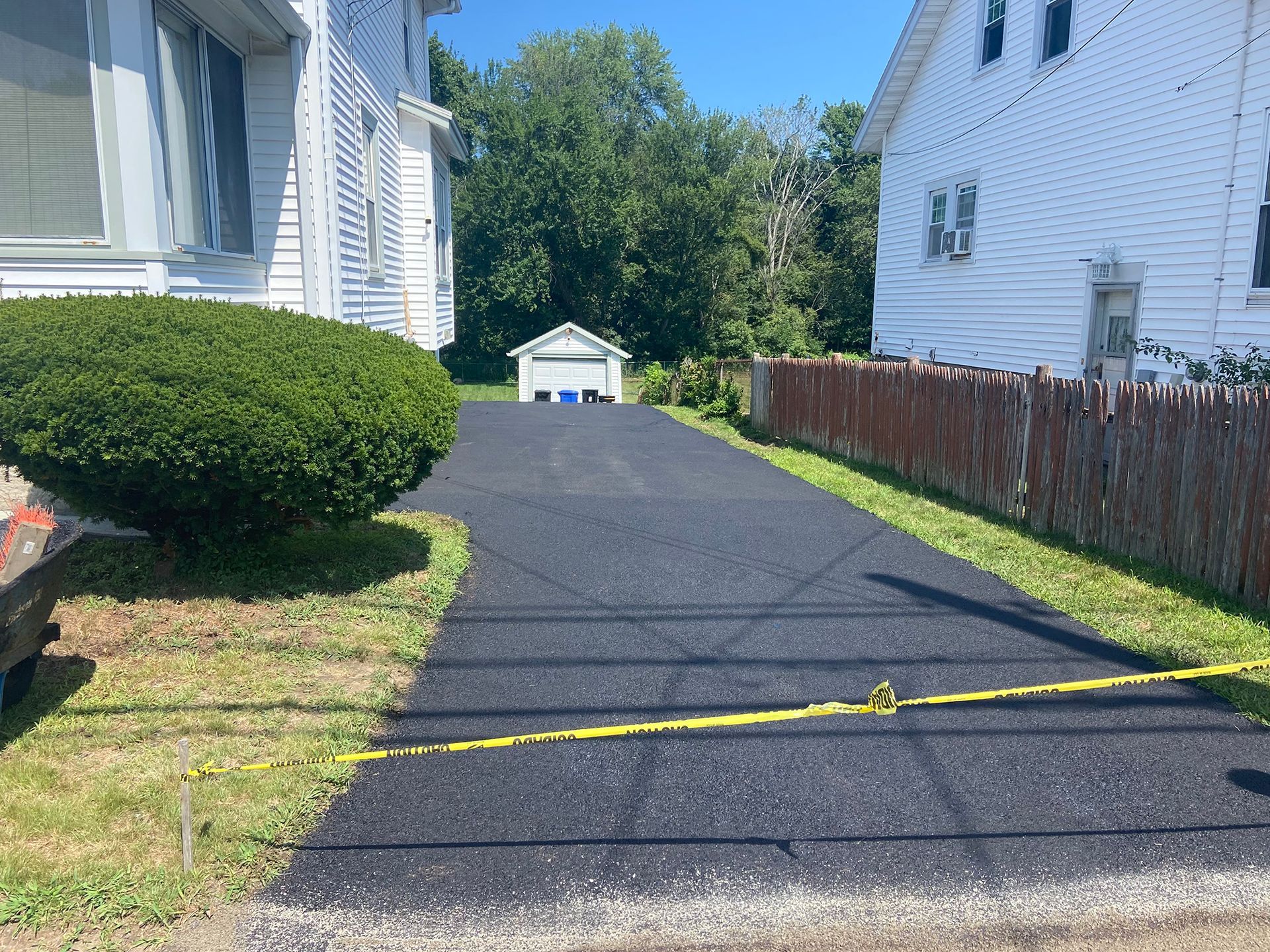 A newly paved black asphalt driveway leads to a small white garage between two residential houses on a sunny day.