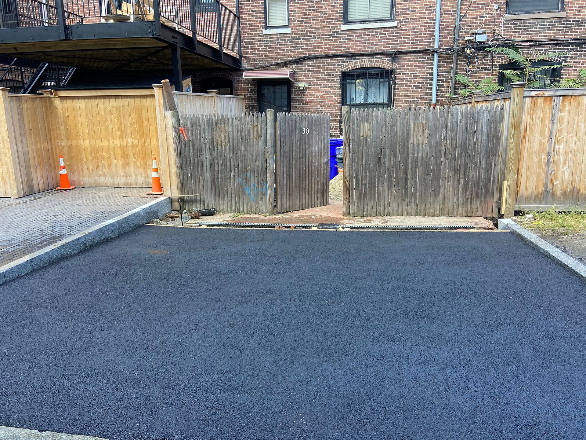 A newly paved asphalt driveway leads to a wooden fence and a brick building with windows in a residential setting.
