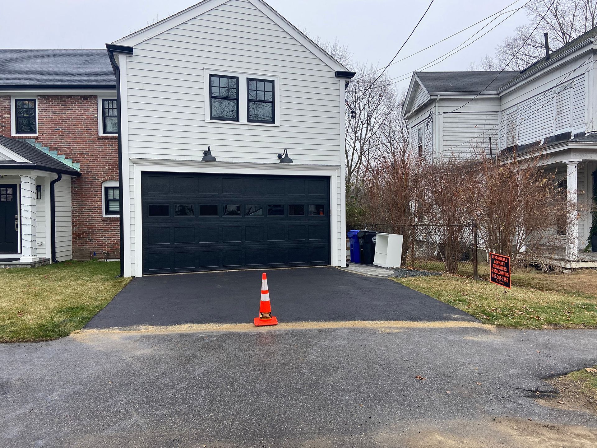 A black-and-white residential garage with a dark door, a freshly paved driveway, and an orange traffic cone in front.