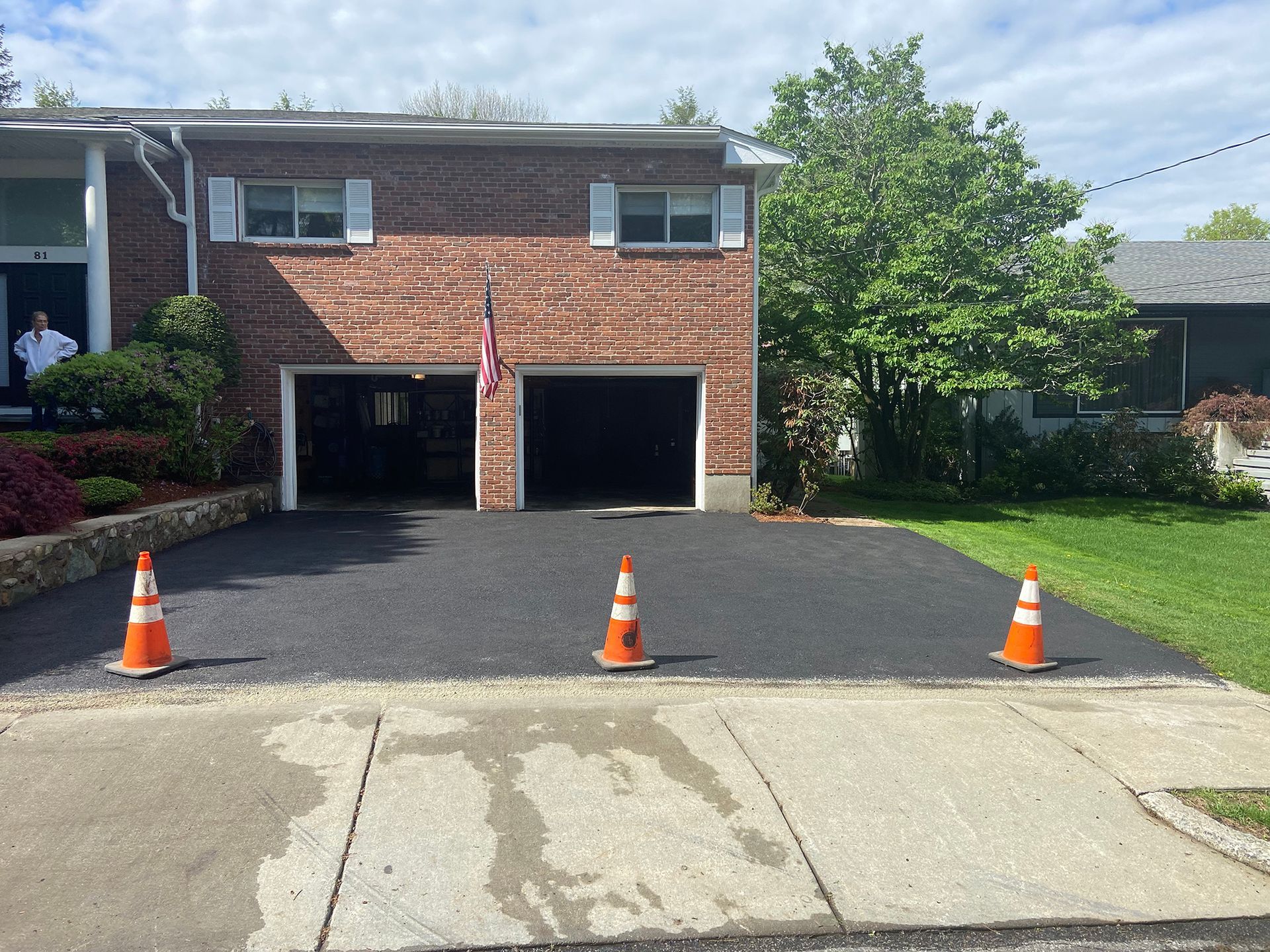 A newly paved asphalt driveway in front of a brick house with two open garage doors and three orange traffic cones.