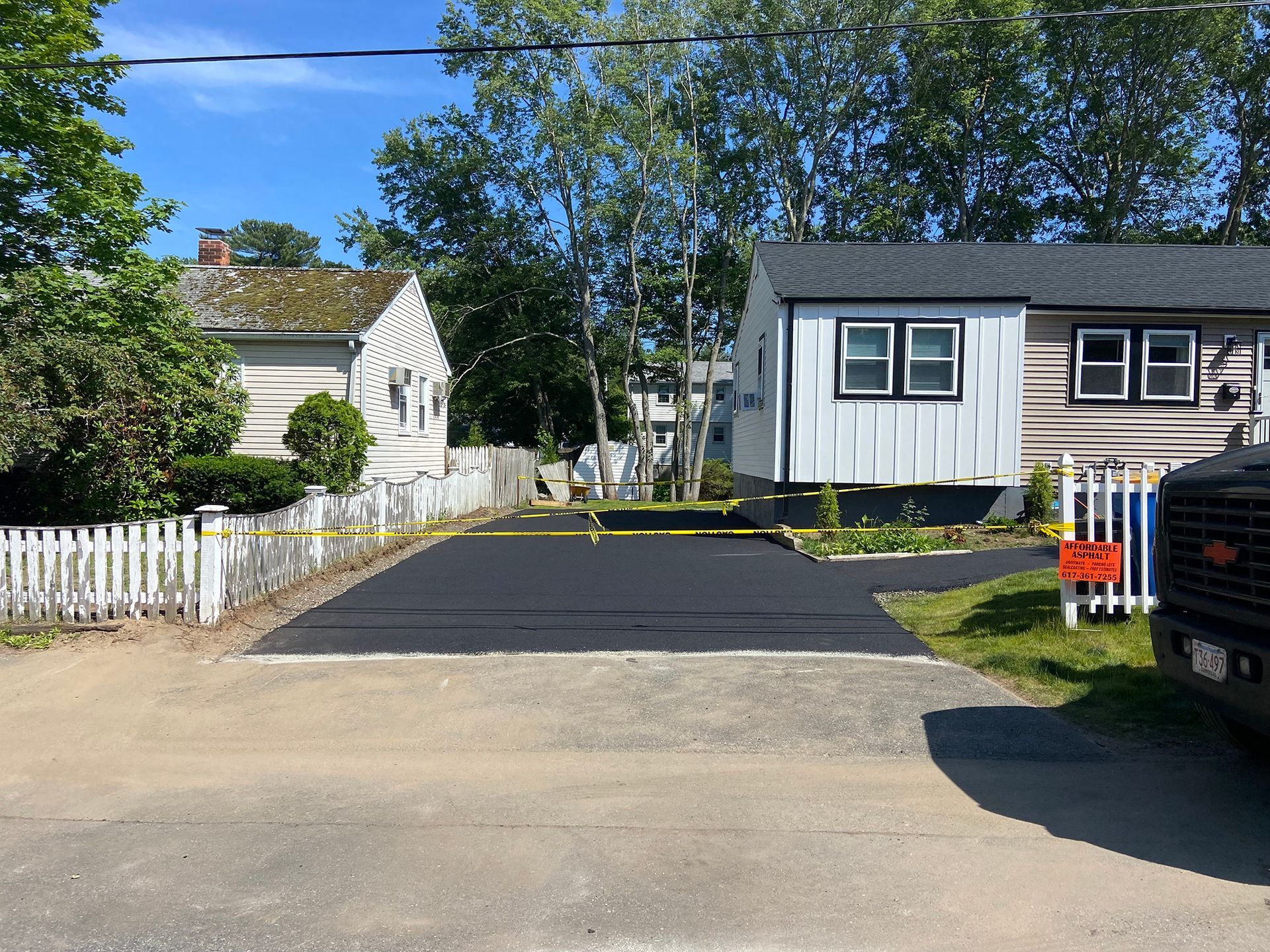 A newly paved asphalt driveway sits between two single-story homes with white picket fences under a bright blue sky.