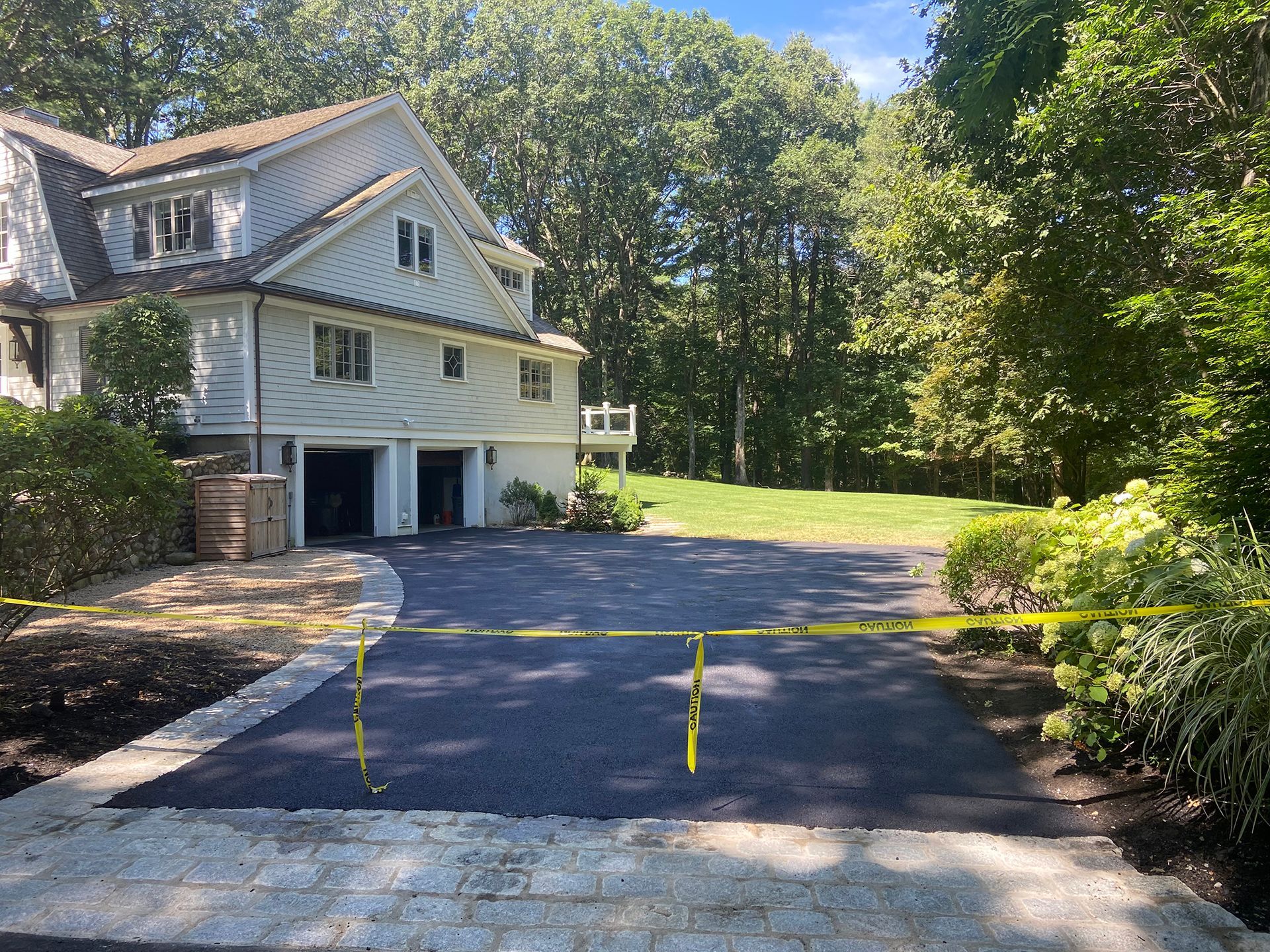 A freshly paved asphalt driveway leads to a white house with a stone apron and yellow caution tape across the front.