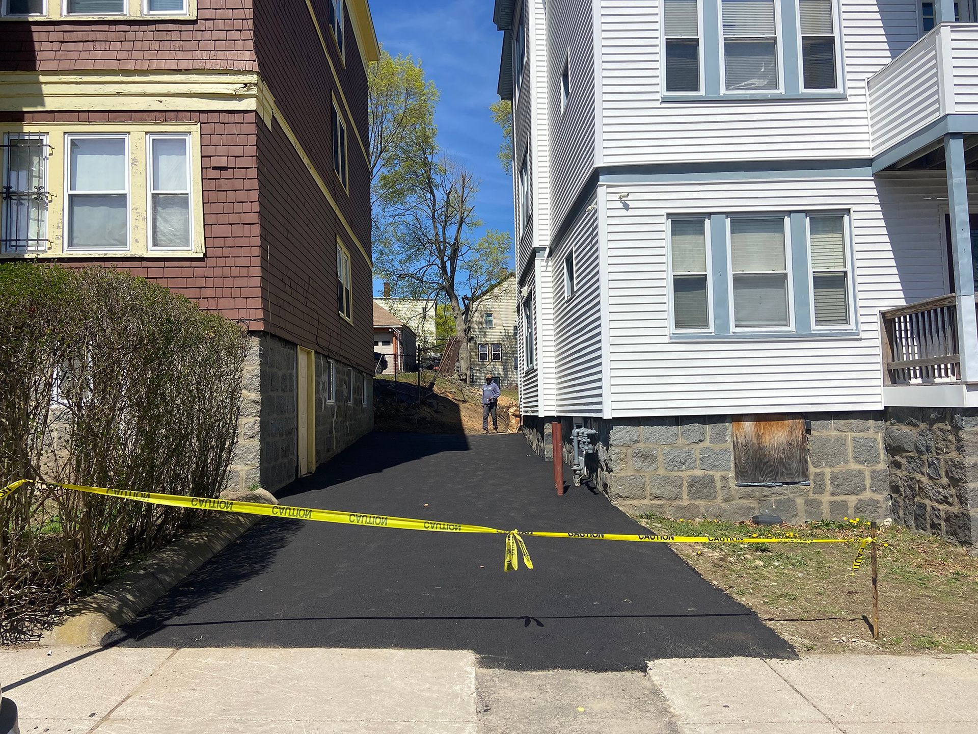 A freshly paved asphalt driveway between two apartment buildings, cordoned off by yellow caution tape.