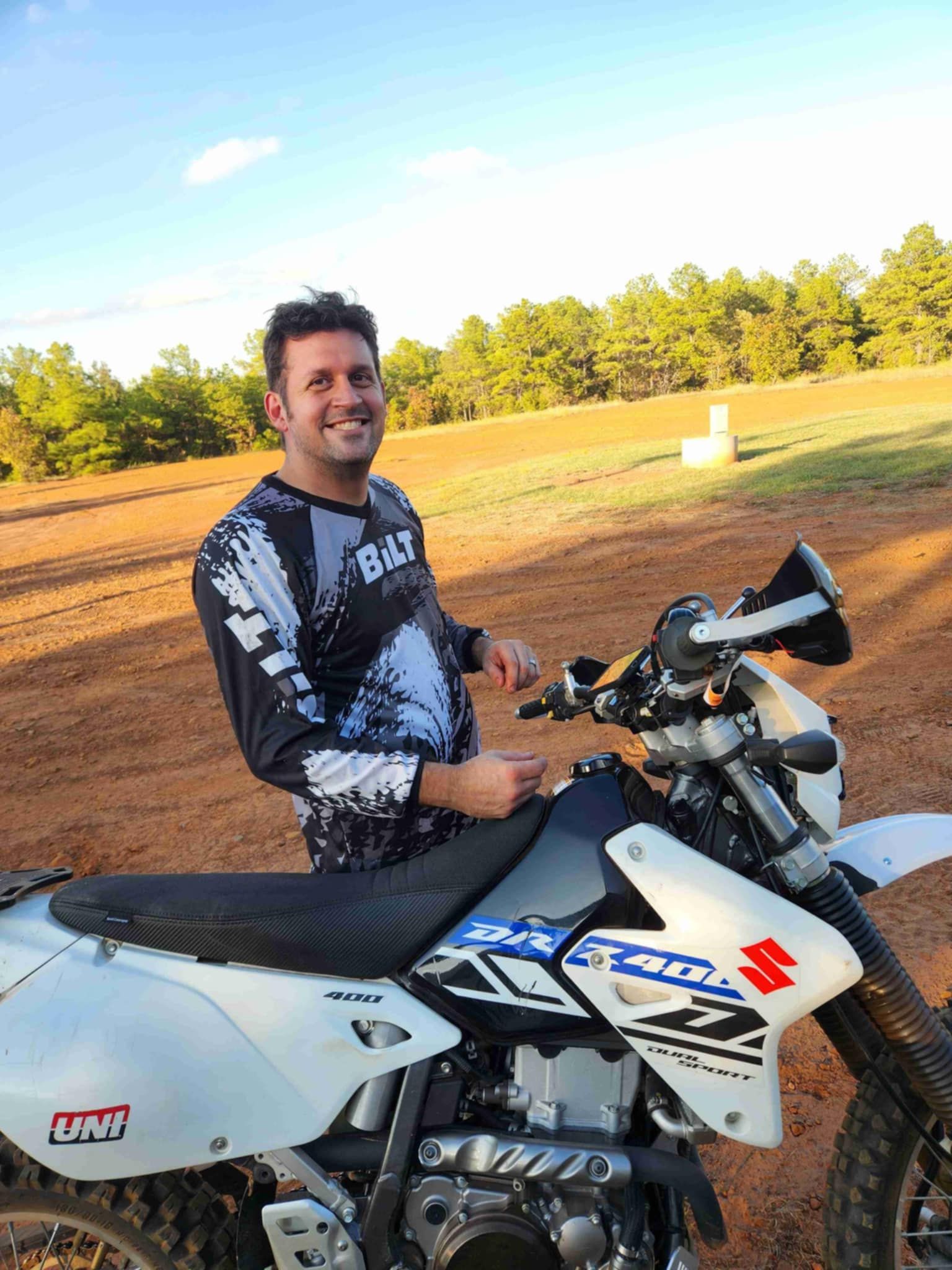 Man in motorcycle gear smiles next to a white and blue Suzuki DRZ400SM dirt bike on a dirt track.