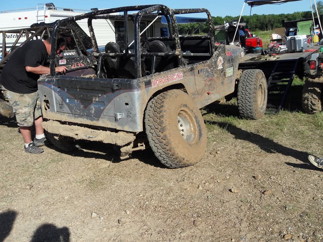 A muddy off-road vehicle being worked on by a person. The scene is outdoors, likely at an off-road event.
