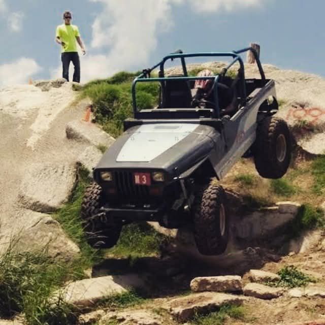 Gray off-road vehicle jumping over rocks on a hillside; a person stands on the rocks watching.