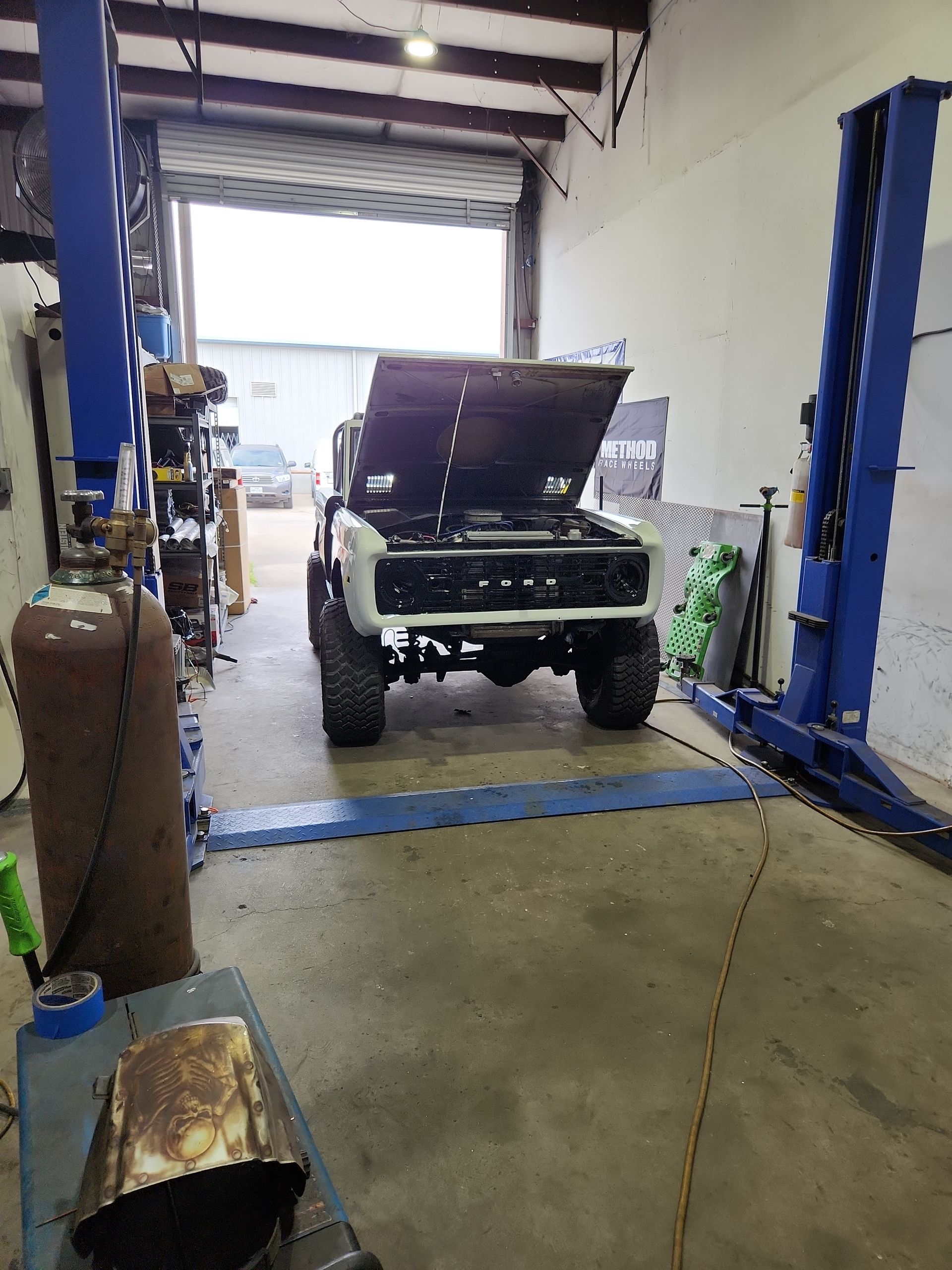 An old white truck with its hood up is inside a garage with the door open. It sits on a lift.